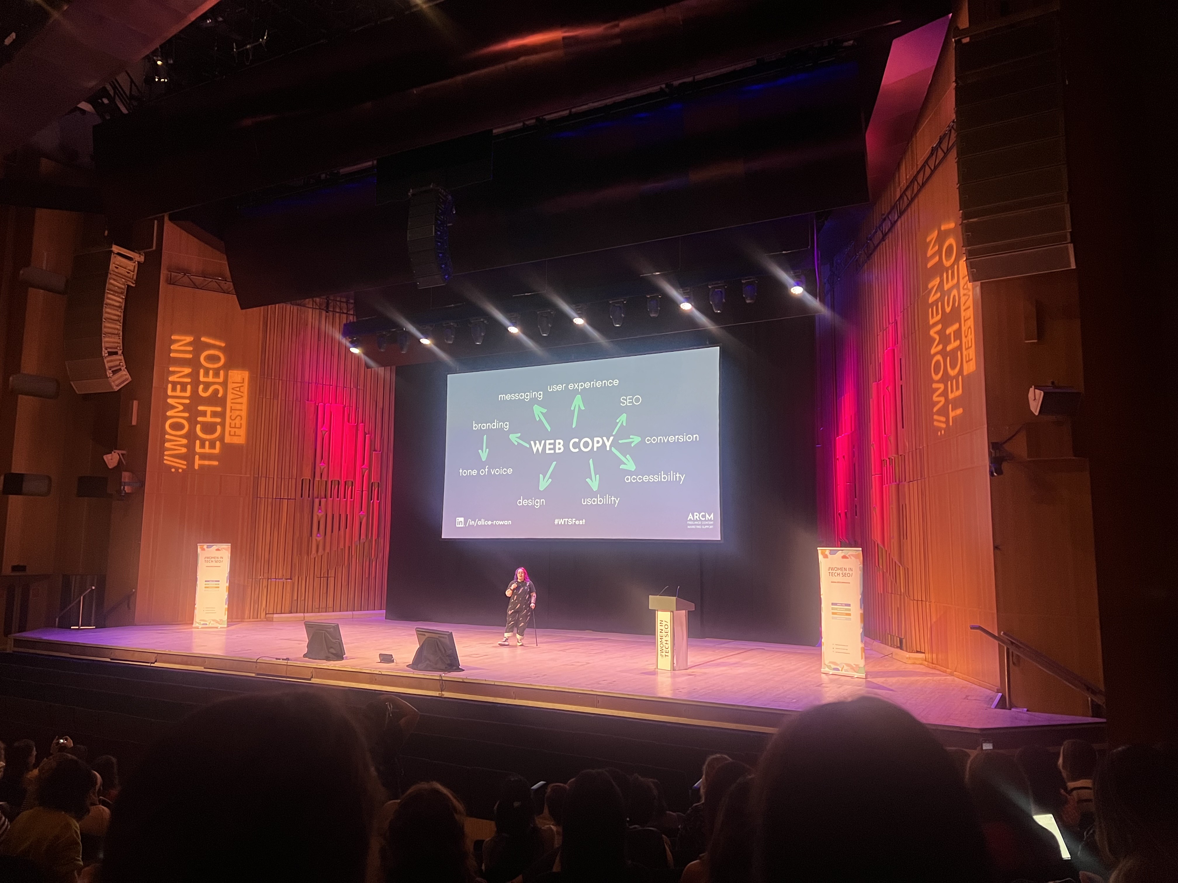 Alice Rowan speaking onstage with a visualisation of web copy on the screen onstage